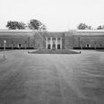 Aarhus Stadion set fra Stadion Allé med John Stampes Plads foran. Billedet er fra efter 1934, da den sidste af de seks statuer langs facaden først opsættes 6. juli 1934, og før marts 1943, hvor den østlige stadionhal nedbrænder. På billedet ses fra venstre langs facaden statuerne Aarhus-pigen eller (Vågnende Kvinde), Uffe hin Spage, Diskoskaster, Manden, Svømmepigen, Atalante, samt i midten Skraberen. Foto: Stadsingeniørens Kontor, Aarhus Stadsarkiv.