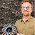 Lektor Søren Munch Kristiansen fra Institut for Geoscience på Aarhus Universitet holder prototypen, som han var med til at udvikle. Bag ham hænger en tavle med et udsnit af udgravningen af køkkenmøddingen ved Vojel Vig på sydkysten af Fur. Foto: David Stott / Journal of Archaeological Science/Peter F. Gammelby, AU